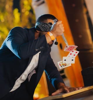 A blindfolded magician in a navy suit throws playing cards into the air at a table.