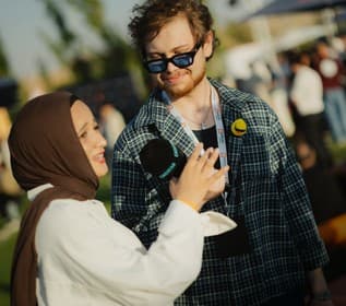 A creator in a hijab interviews a guest in a checked shirt with a handheld microphone on the lawn.