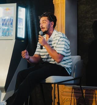 A speaker in a striped shirt sits on stage with a microphone, mid-sentence.