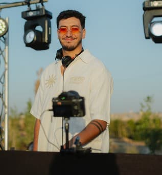 A DJ in orange sunglasses and headphones behind the decks under stage lights.