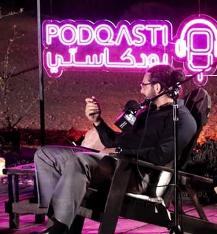 A host records a podcast at night in front of a pink neon Podqasti sign.